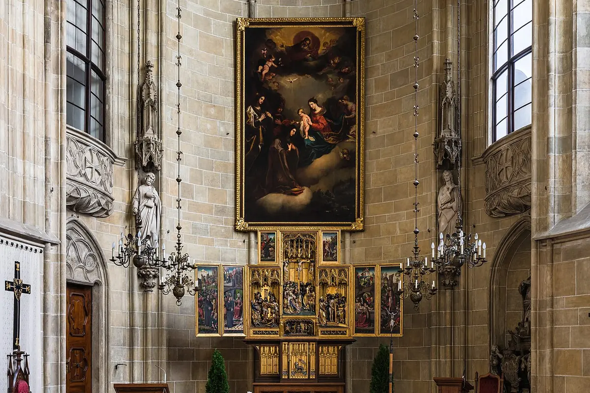 Prezbiterium kościoła Deutschordenskirche w Wiedniu: gotyckie sklepienie gwiaździste, wysokie okna, duży obraz ołtarzowy nad złoconym skrzydłowym ołtarzem i rzędy drewnianych ławek.