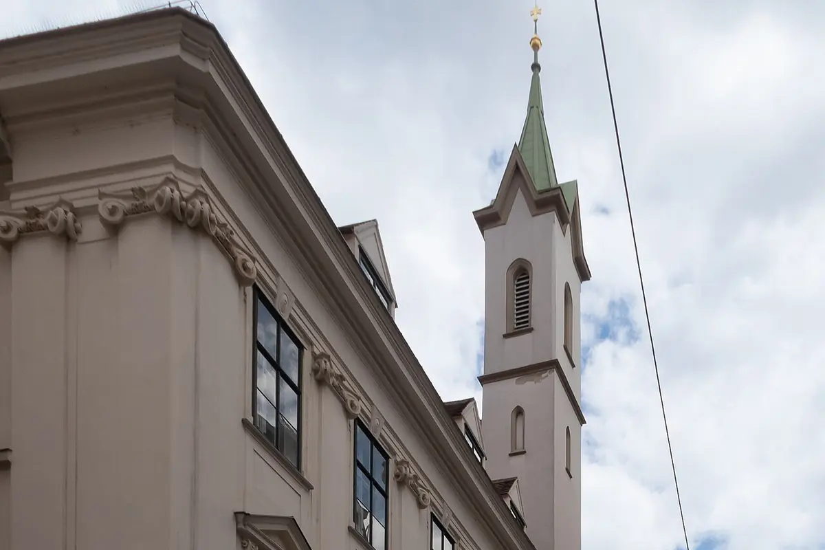 Barokowy narożnik domu Zakonu Krzyżackiego przy Singerstraße w Wiedniu, a za linią dachu wznosi się smukła wieża kościelna zwieńczona złotym krzyżem.